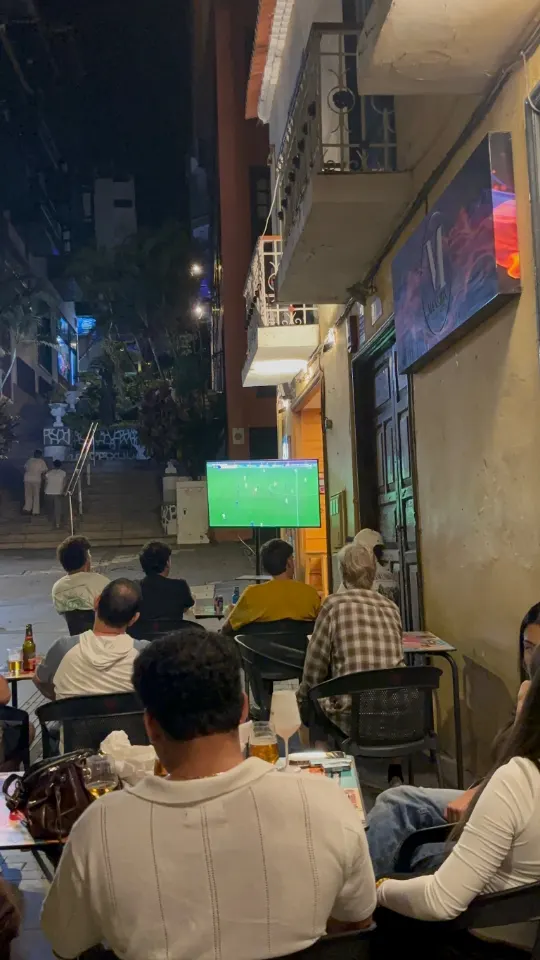 Clientes en la terraza de noche viendo el fútbol en la pantalla exterior
